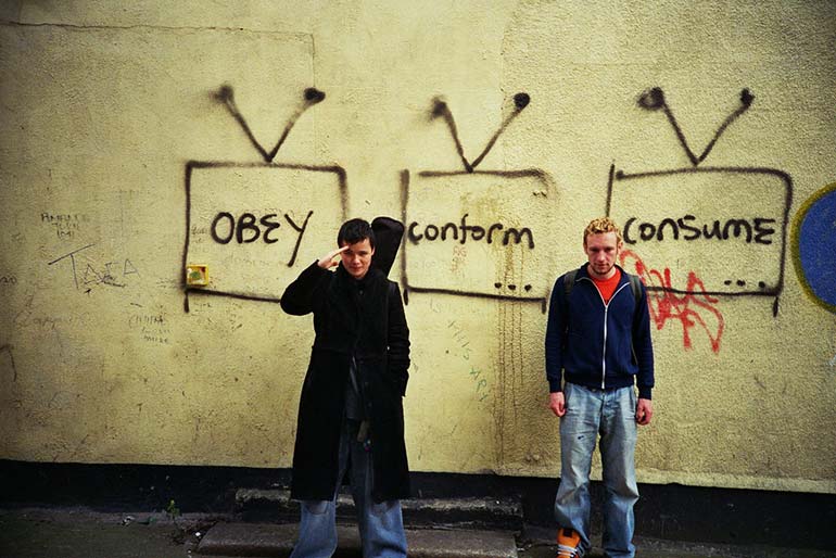 Two adolescent males standing in front of a wall graffitied with the words "Obey", "Conform", and "Consume".