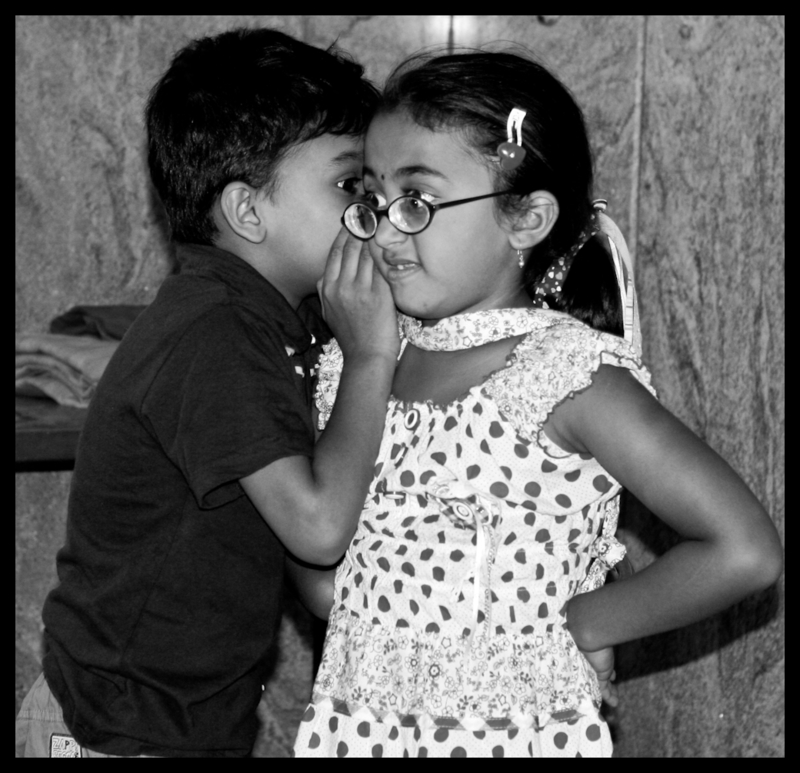Small boy whispering in the ear of a young girl, who has a look of shock and disgust on her face.