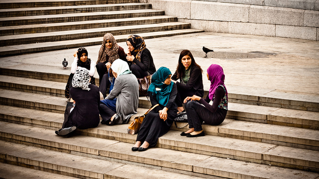 Two groups of teenage girls, most of whom are wearing head scarves, sitting and chatting on some steps.
