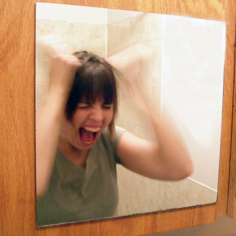 A woman stands in front of a mirror screaming and pulling her own hair.