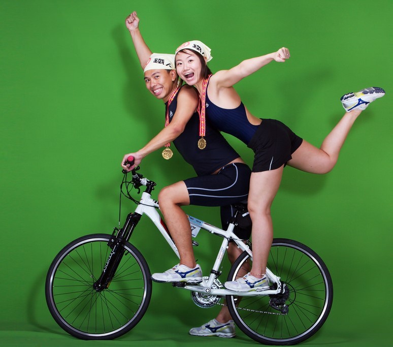 An young, athletic couple pose for photos on their bike while wearing medals they've won in competition.