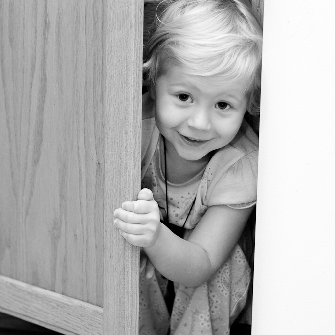 A young girl smiles as she peeks out from a hiding place.
