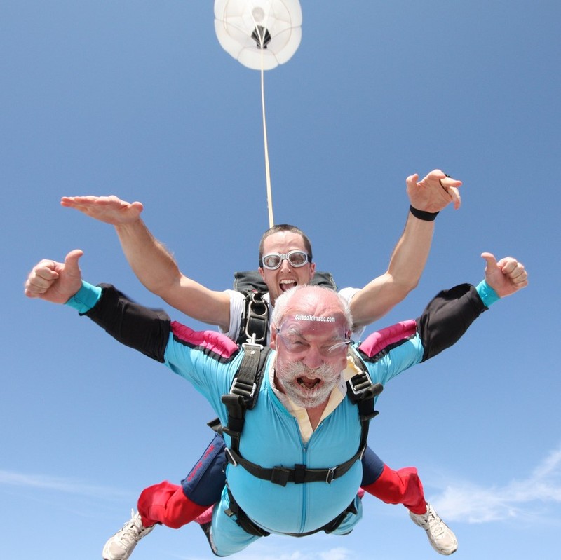 An older man with white hair and beard skydives in tandem with a younger jumpmaster.