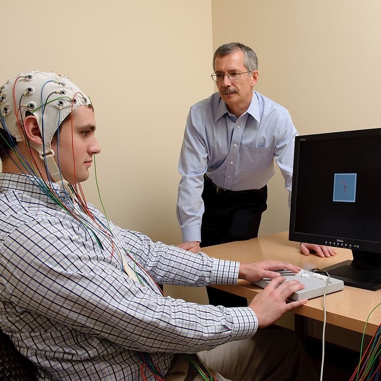 A study participant wears an EEG cap and uses a touch pad to react to images on a computer screen. An experimenter stands by observing.