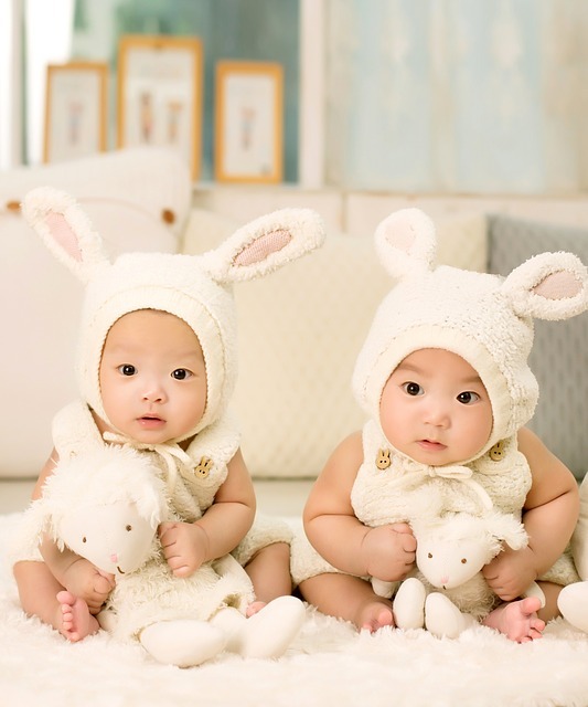 Twin boys sit together dressed in matching clothes and hats and holding similar stuffed animals.