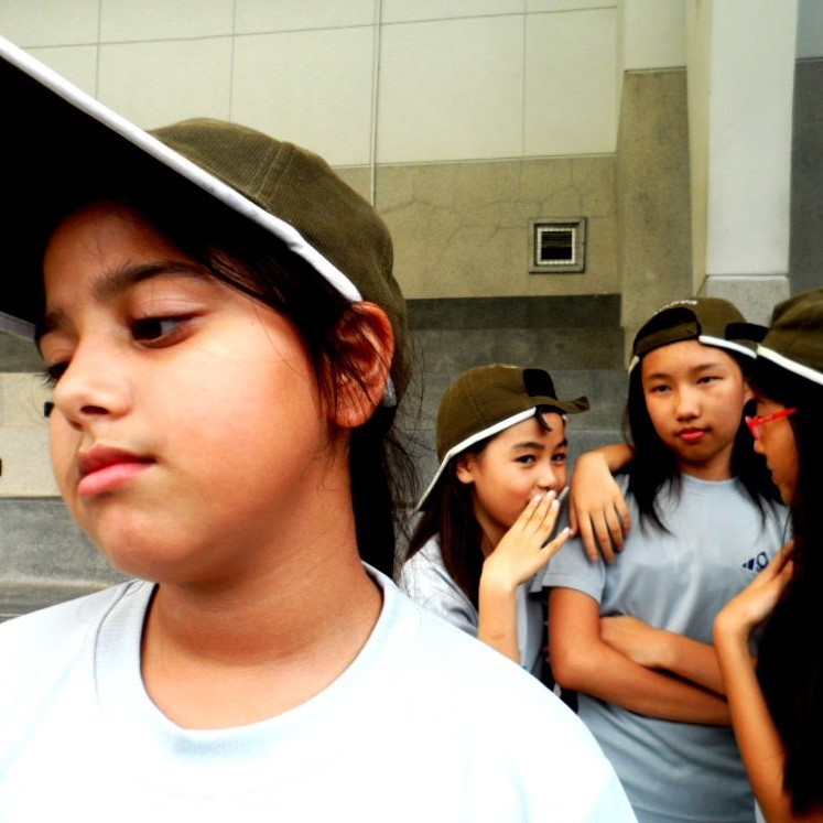 A sad looking girl stands in the foreground as a group of her classmates stands behind looking at her and whispering behind their hands.