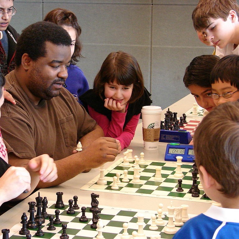 A group of children standby watching an adult playing a game of chess.