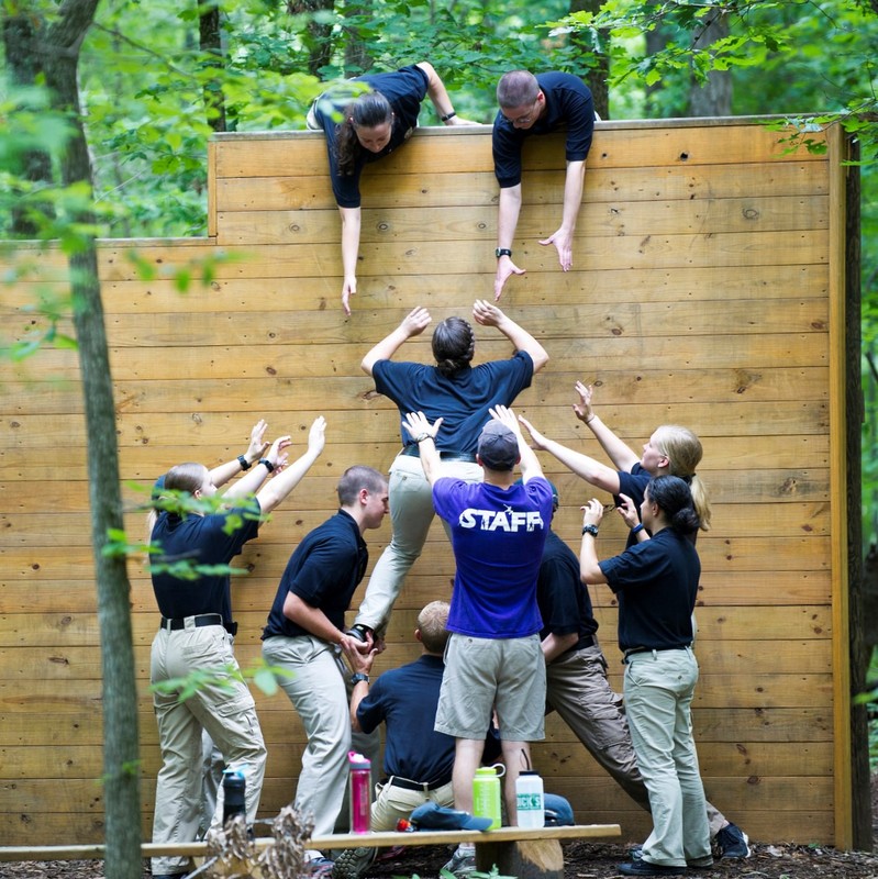 A team of people on an obstacle course work together to boost one member of the group over a high wall.