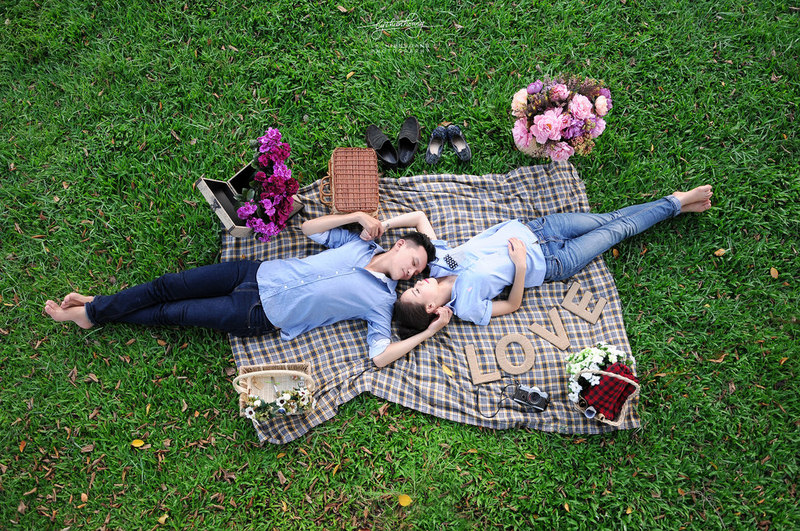 A romantic couple lying on a picnic blanket surrounded by bouquets of flowers.