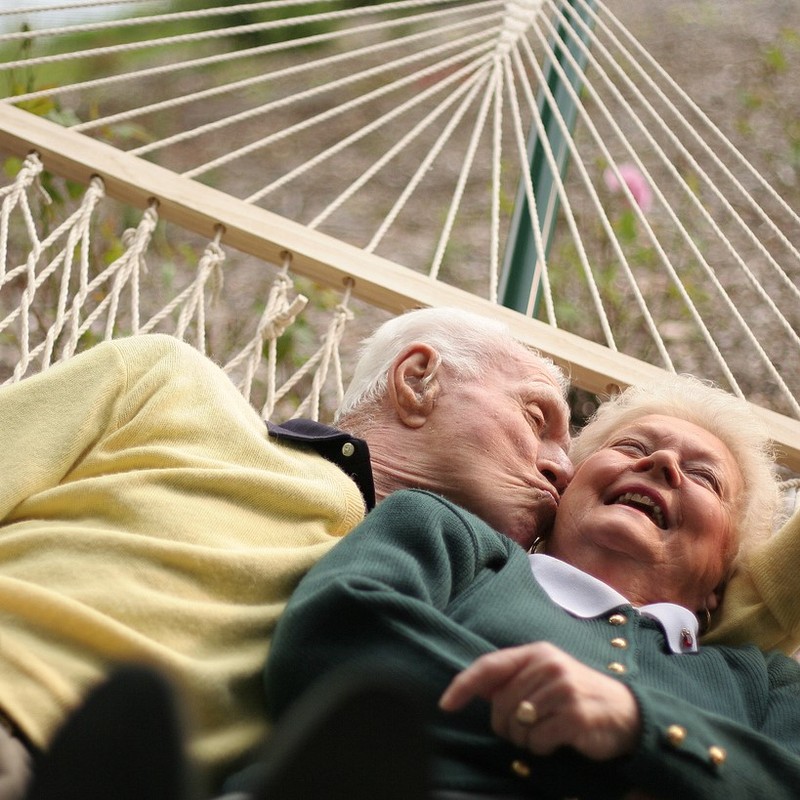 An elderly couple lie together in a hammock. The man kisses the woman on the cheek as she laughs.