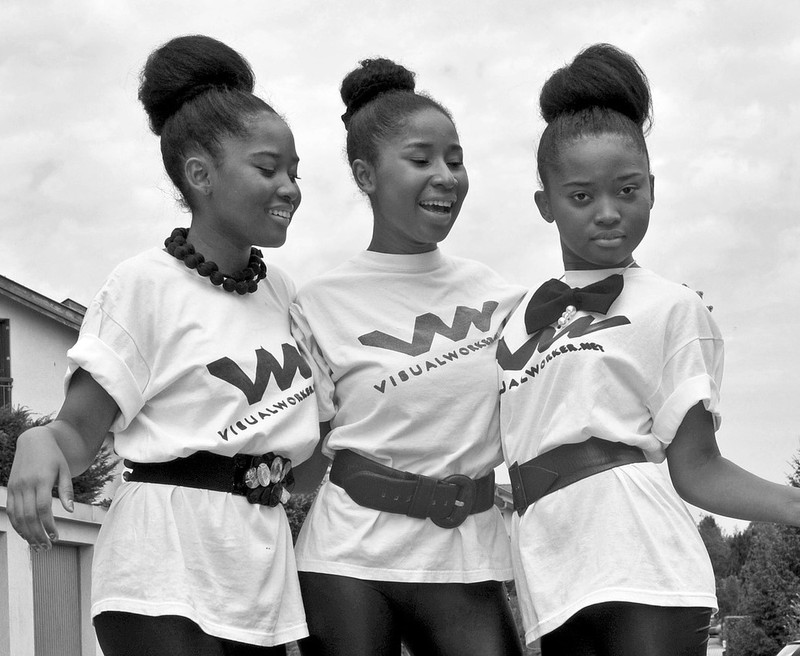 A group of teen girls with matching shirts and hair styles pose together with their arms around one another.