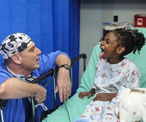 A doctor visits a young patient in a hospital bed. She playfully sticks her tongue out to show the doctor.
