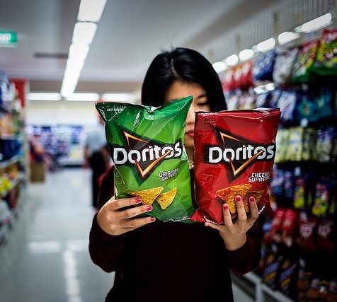 A woman in a supermarket aisle holds two different types of Doritos and carefully reads the packages.