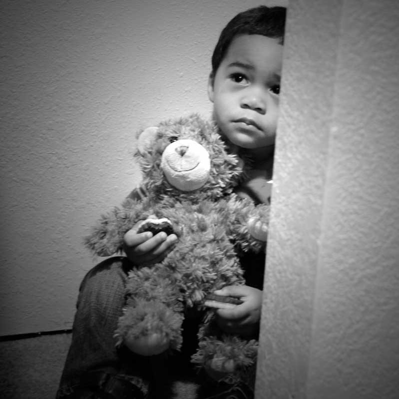An unhappy looking little boy sits with his teddy bear on the floor of a closet.