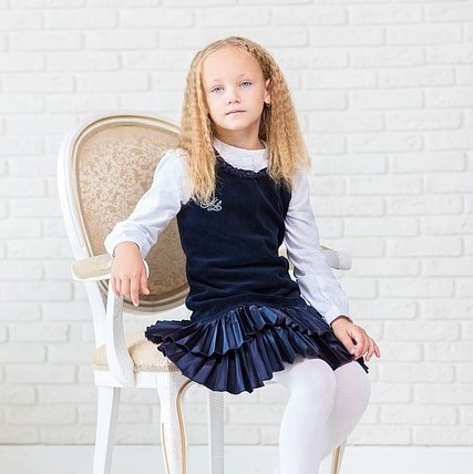 A young girl poses for a portrait dressed in an outfit that society might generally considered appropriately feminine - white tights, pleated velvet skirt, ruffled collar, and tailored cuff.