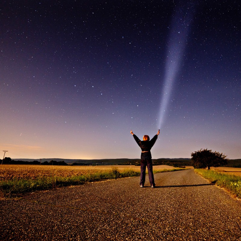 A woman stands in the middle of a country road at night and reaches toward the star-filled sky above.