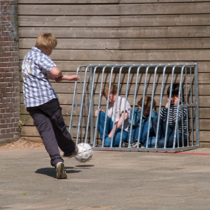 A playground bully kicks a soccer ball at a group of students huddled behind a barrier.