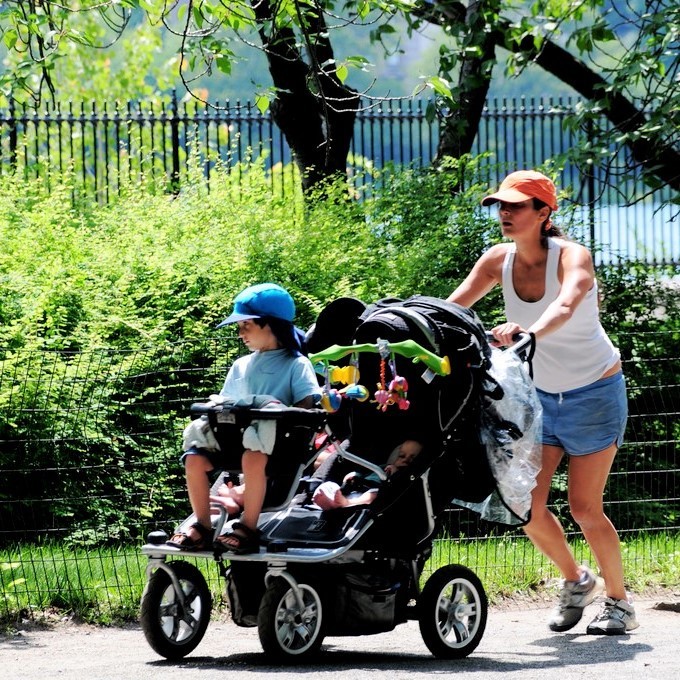 A woman jogs through a park with three young children in a stroller.