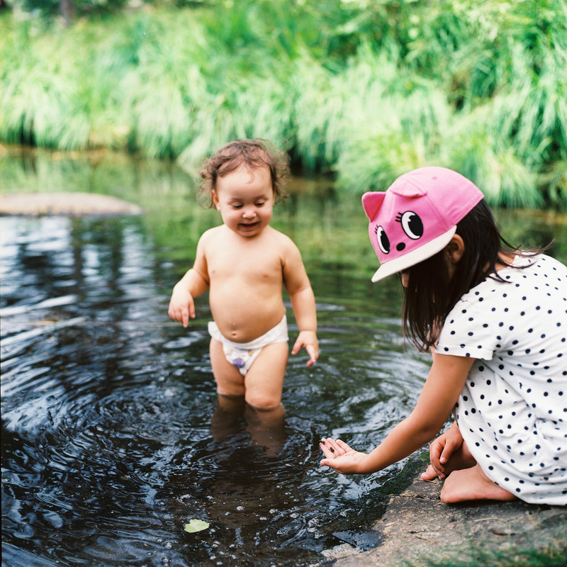 A toddler in a diaper wades in a pond while his sister picks up pebbles on the bank.
