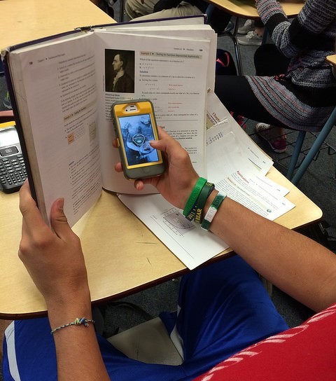 A student sits at a desk using a cell phone hidden behind a textbook.