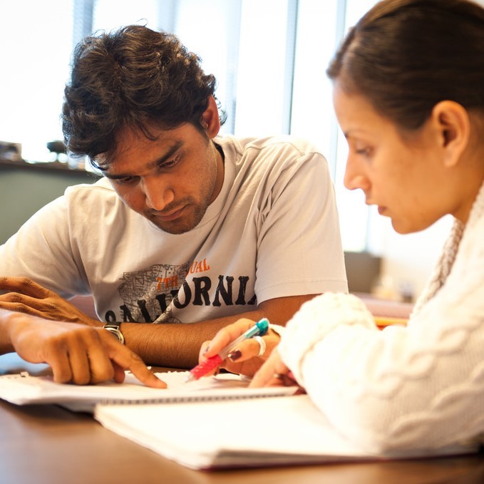 A male and female student work together at a table and focus on details in a notebook in front of them. 