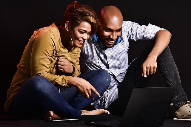 A young couple smiling and watching something on their laptop screen, the woman is pointing something out to the man.