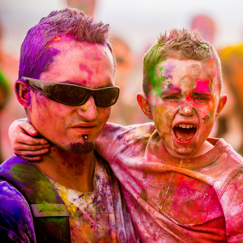 Un padre y un hijo sonríen y gritan después de terminar una emocionante carrera llamada The Color Run. Ambos están cubiertos de pies a cabeza en muchos tonos de polvo de colores brillantes.