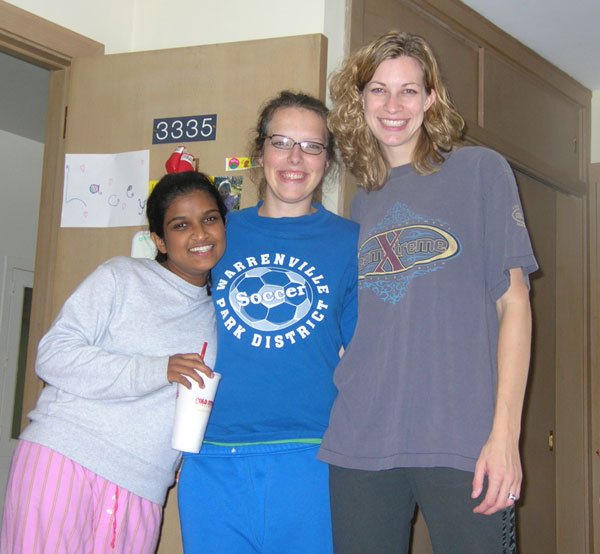 Drei College-Freunde stehen zusammen in einem Wohnheimzimmer. Die Frau auf der rechten Seite ist 15 Zentimeter größer als die Frau in der Mitte. Die Frau in der Mitte ist 15 cm größer als die Frau auf der linken Seite.
