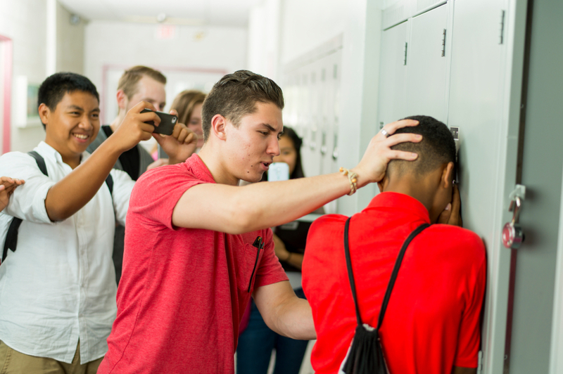 A teenage boy pushes another boy up against a school locker as a group of fellow students watch.