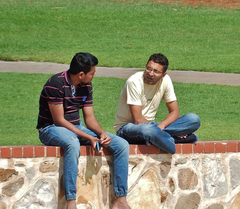 Two young men sit together outside on a sunny day having a conversation.