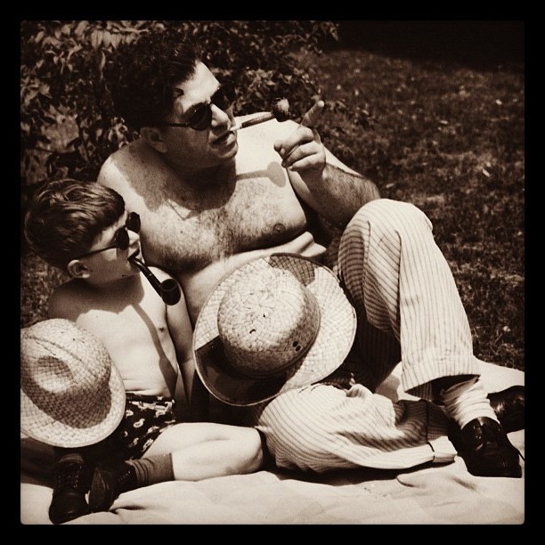A father and his young son sit together on a blanket on the lawn on a sunny day. Each have their shirts removed and are dressed almost identically including straw hats, sunglasses, and pipes.