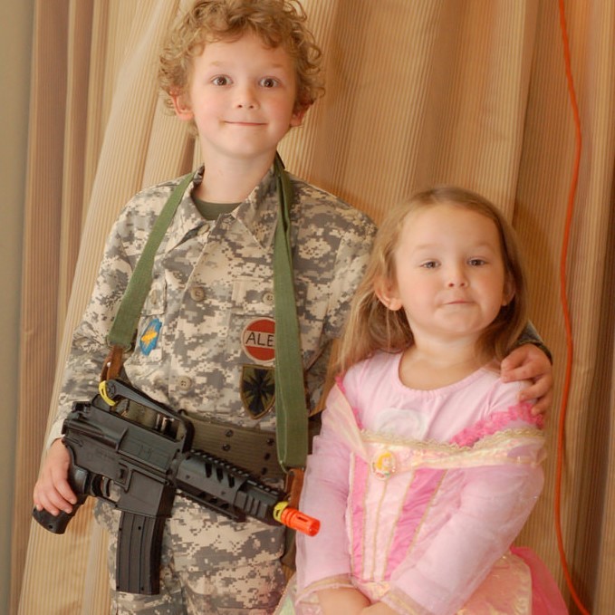 Un hermano y una hermana están uno al lado del otro. Está vestido con un uniforme militar de camuflaje y sostiene una pistola de juguete. Ella está vestida con un bonito vestido de princesa rosa.