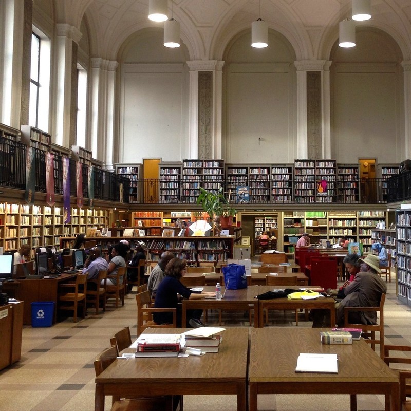 Cajas altas de libros rodean un área central llena de escritorios mientras la gente se sienta leyendo y accediendo a la información desde computadoras en una biblioteca pública.