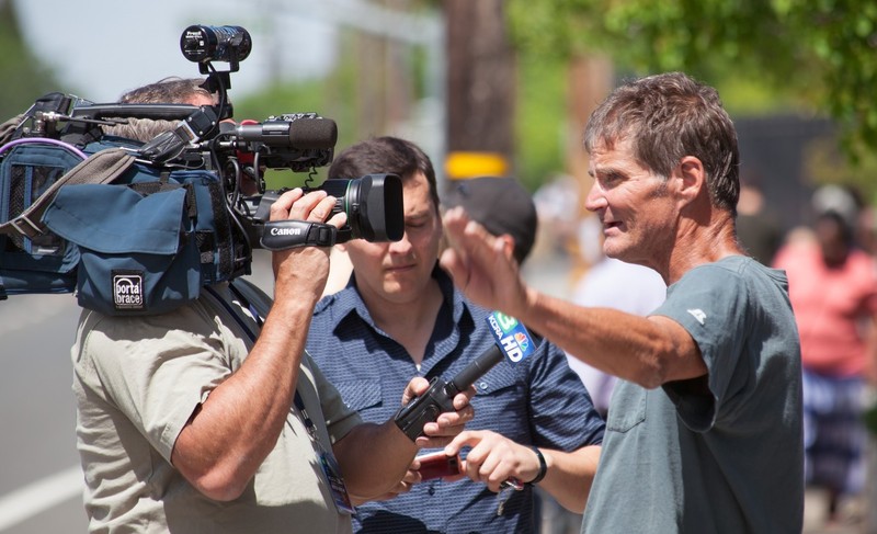 A man stands in front of a television camera and microphone as he describes an event he has witnessed. 