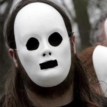 A man is seen wearing an expressionless white plaster mask with black eyes and mouth.