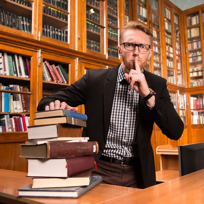 Un hombre vestido con chamarra y pajarita se encuentra detrás de un escritorio apilado con libros. Tiene una mirada severa en su rostro y se está metiendo un dedo en los labios solicitando silencio. Detrás del hombre hay estantes forrados con libros.