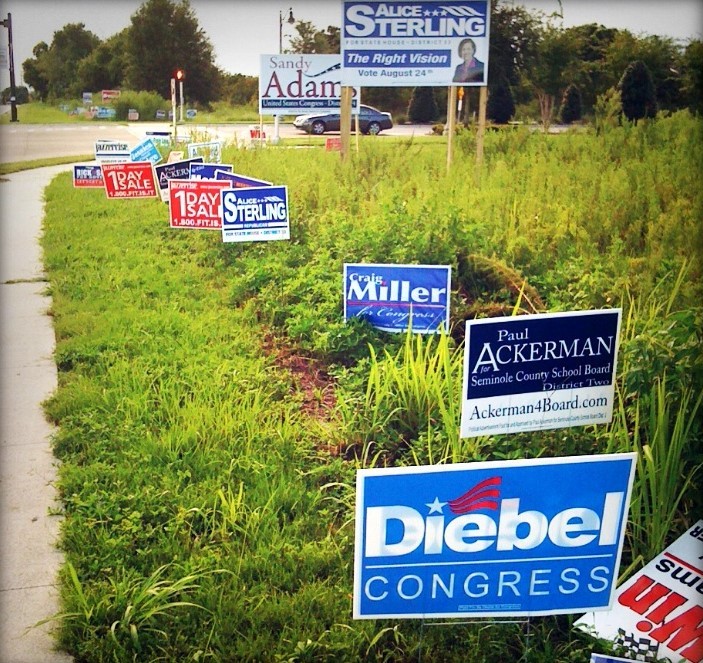 Un campo cerca de una carretera está lleno de letreros de campaña política y anuncios para una “venta de 1 día”.