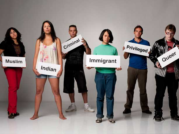 A group of people stand holding signs labeling them as others perceive them. The signs include, &quot;Muslim&quot;, &quot;Indian&quot;, &quot;Criminal&quot;, &quot;Immigrant&quot;, &quot;Privileged&quot;, and &quot;Queer.&quot;