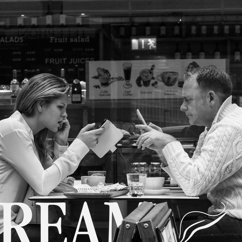 A man and a woman sit across from one another at a small table in a coffee shop. Both of them are staring at their own smartphone rather than engaging with each other.