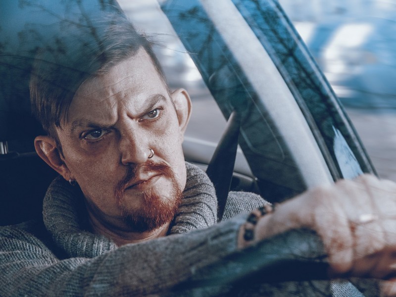A man with an angry expression on his face sits behind the wheel of a car staring straight ahead.