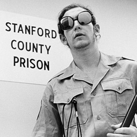 Foto de un guardia participante del experimento de la prisión de Stanford vistiendo gafas de sol y sosteniendo una porra.