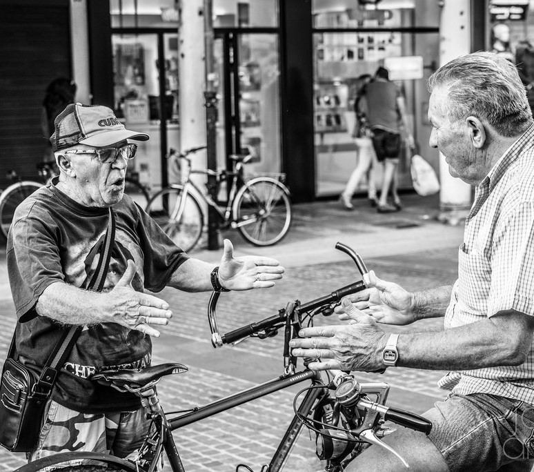 Two men stand talking in the street, each making nearly identical hand gestures.