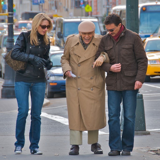 A younger man and woman help an elderly gentleman down the street.
