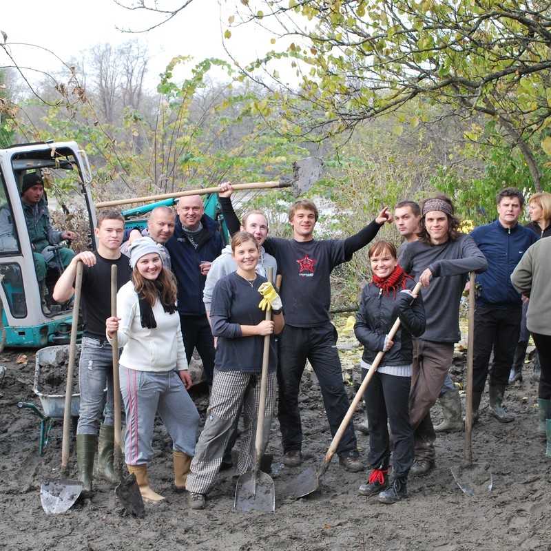 A group of men and women stand together in a muddy field with shovels and wheelbarrows as they participate in an outdoor volunteer project.