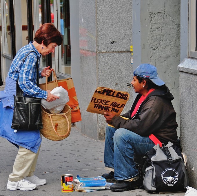 A woman stops on the sidewalk to offer food to a man holding a sign reading "Homeless, please help Thank you."