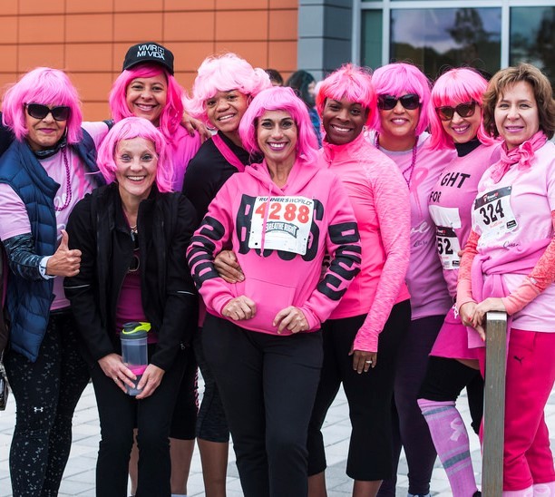 A group of women wearing pink wigs and pink shirts pose together at the conclusion of a 5K race in support of those with breast cancer.