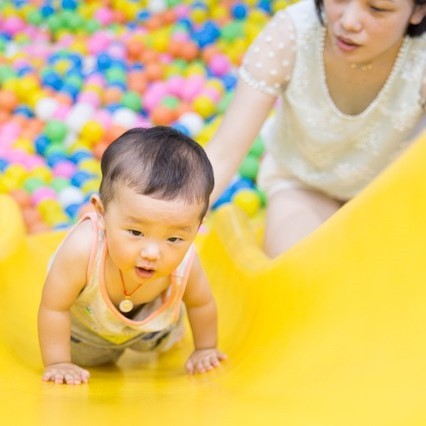 A toddler crawls up a slide in a play area as its mother stands close behind.