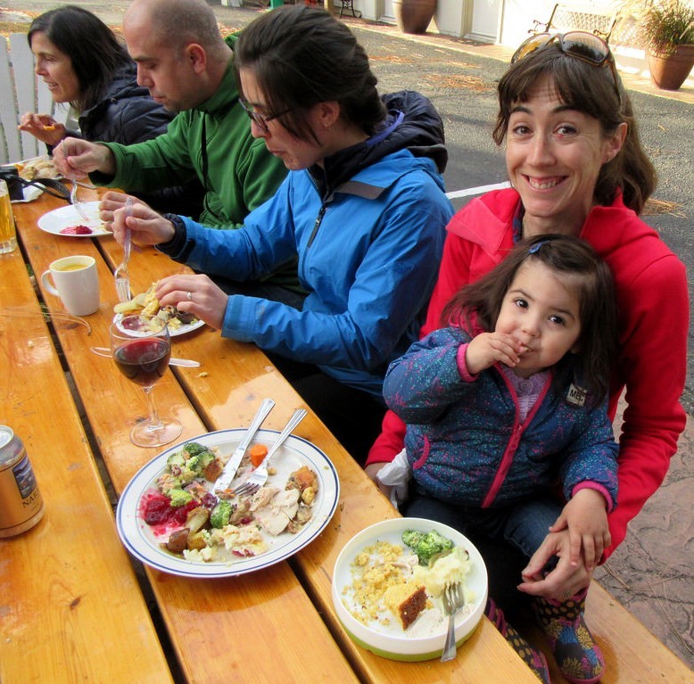 Una joven se sienta en el regazo de su madre durante una comida navideña mientras un grupo de adultos alrededor de la mesa disfruta de comida y bebida.