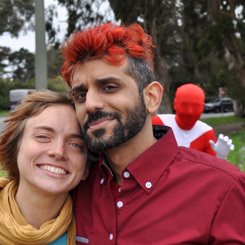A couple pose for a picture in the foreground as a man in a red bodysuit and mask photo bombs them in the distance.