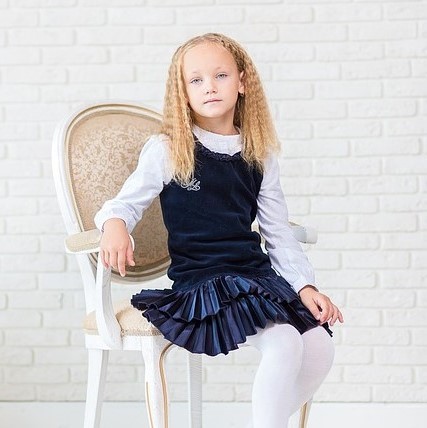 Una joven posa para un retrato vestido con un atuendo que la sociedad generalmente podría considerar apropiadamente femenino: medias blancas, falda plisada de terciopelo, cuello con volantes y puños entallados.